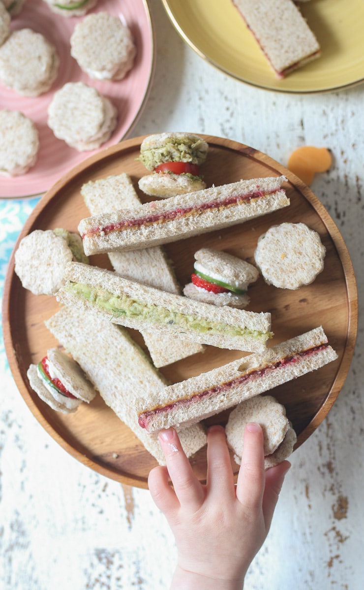 Toddler hand reaching for a finger sandwich on a wooden plate.