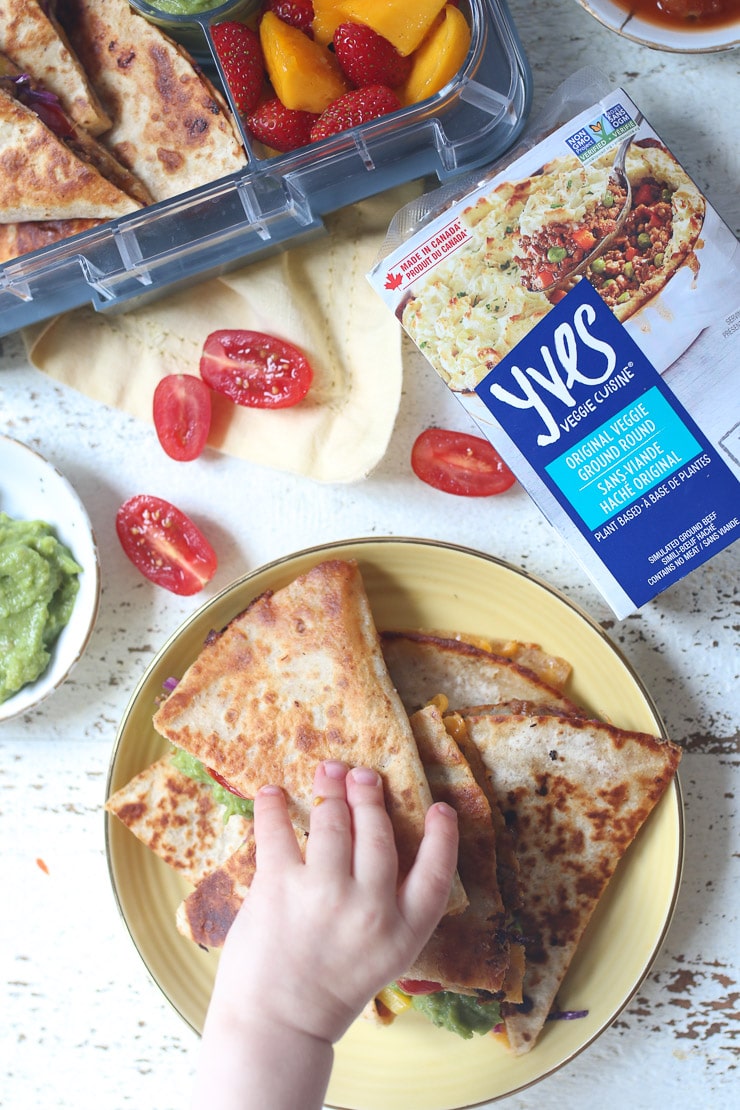 Toddler hand reaching for vegan baked quesadillas from a yellow plate.