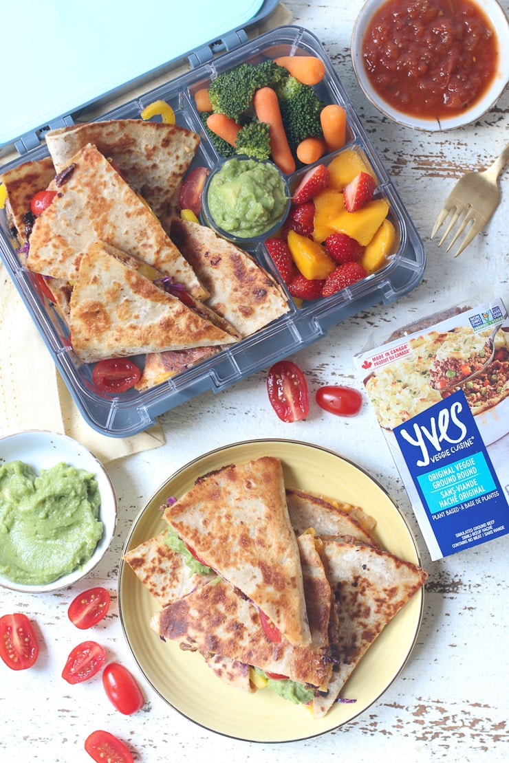 Quesadillas on a yellow plate next to a lunch box with veggies.