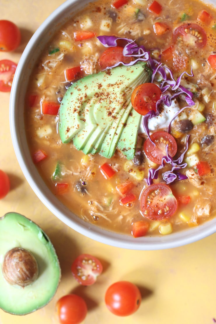 Birds eye view of healthy chicken tortilla soup in white bowl garnished with fresh vegetables and avocado slices on a wooden table sitting next to half of an avocado