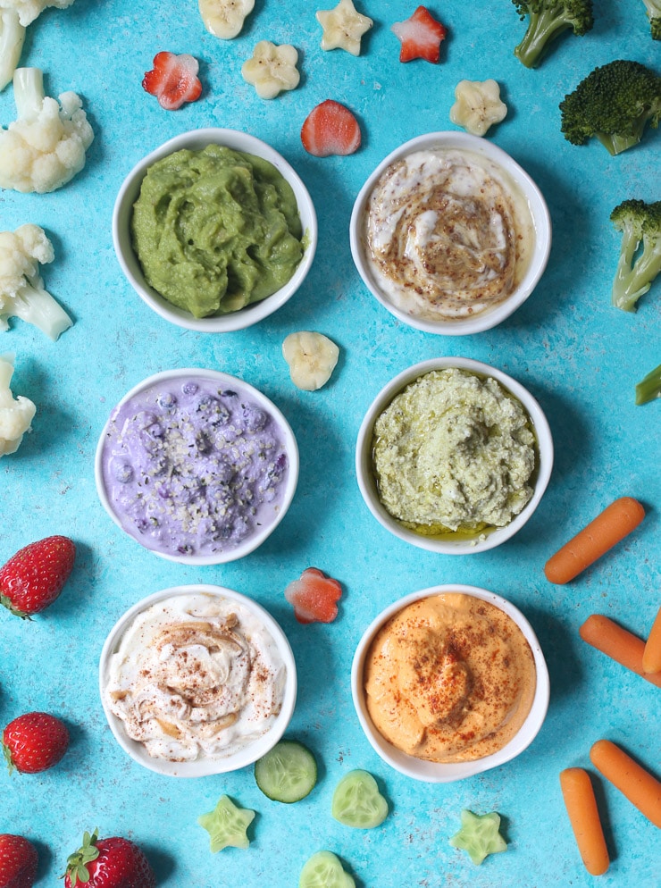 birds eye view of a variety of different dips in small white bowls for toddlers surrounded by fruits and vegetables