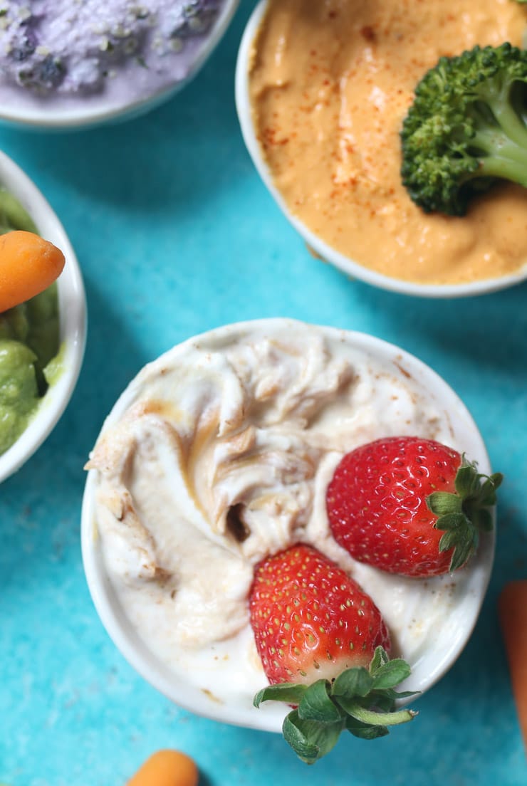 close up image of three varieties of dips for toddlers garnished with fruits and vegetables in small white bowls on a blue background