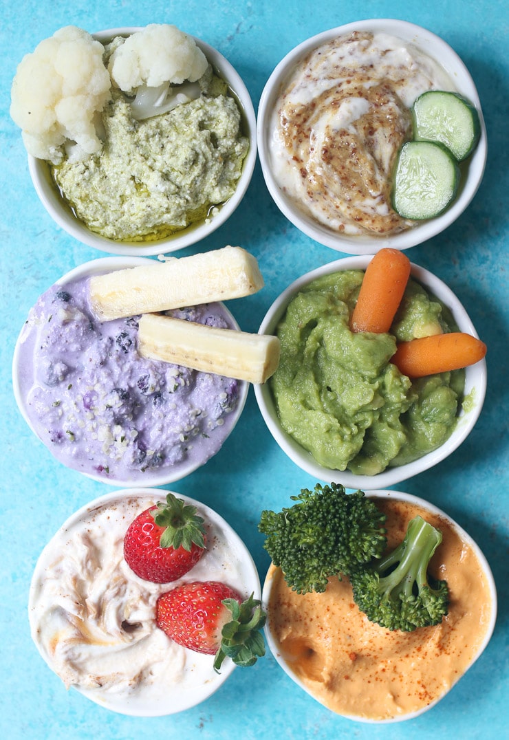 birds eye view of six different dips for toddlers in small white bowls garnished with fruits and vegetables