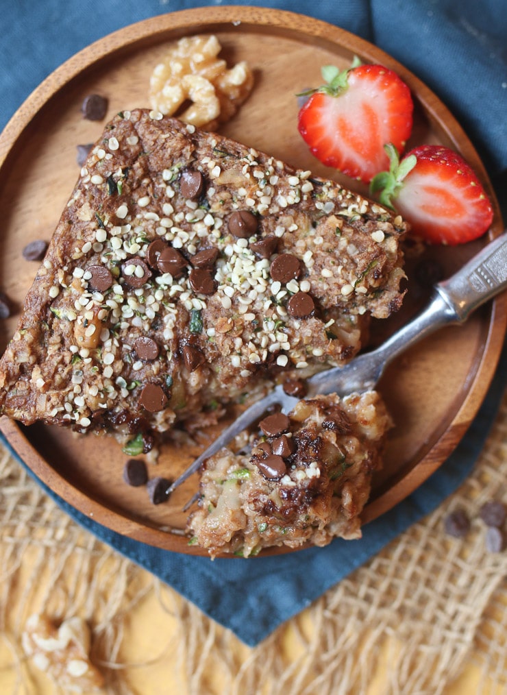 Birds eye view of a piece of chocolate zucchini baked oatmeal square on a wooden plate garnished with hemp seeds and chocolate chips with strawberries on the side 