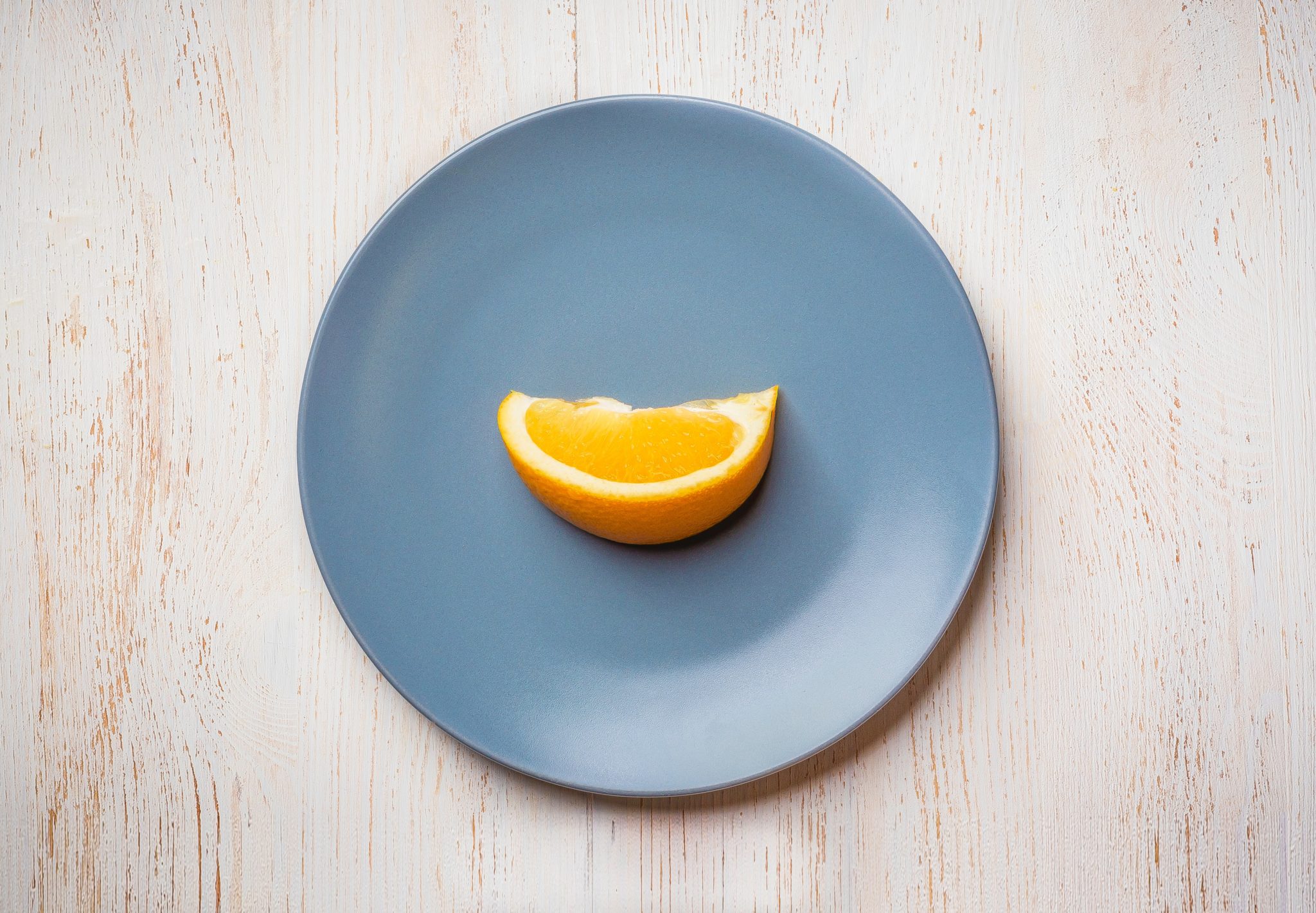 Birds eye view of a blue plate with a single orange slice on it against a light background.