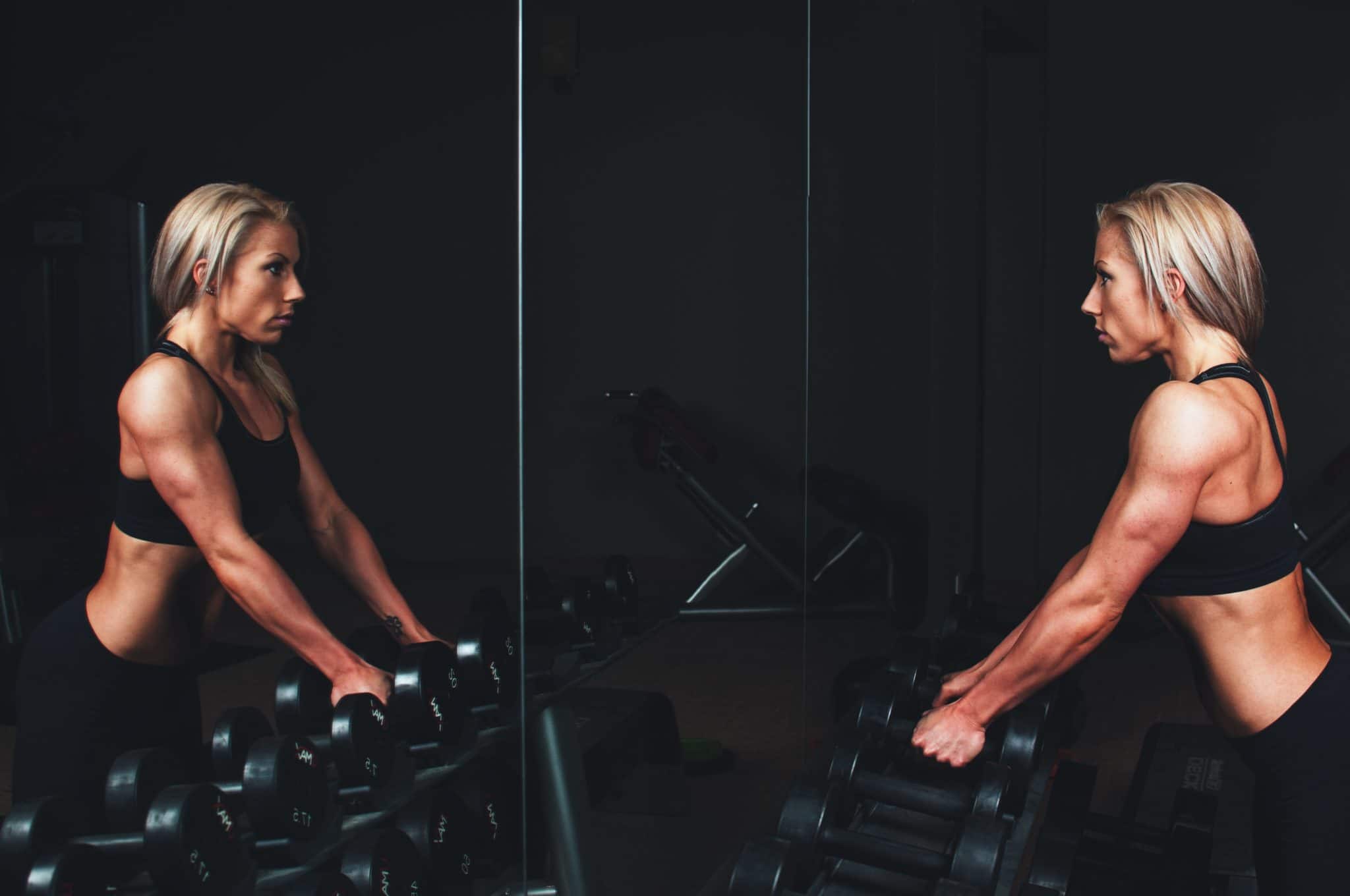 Female body builder standing in front of mirror holding weights.