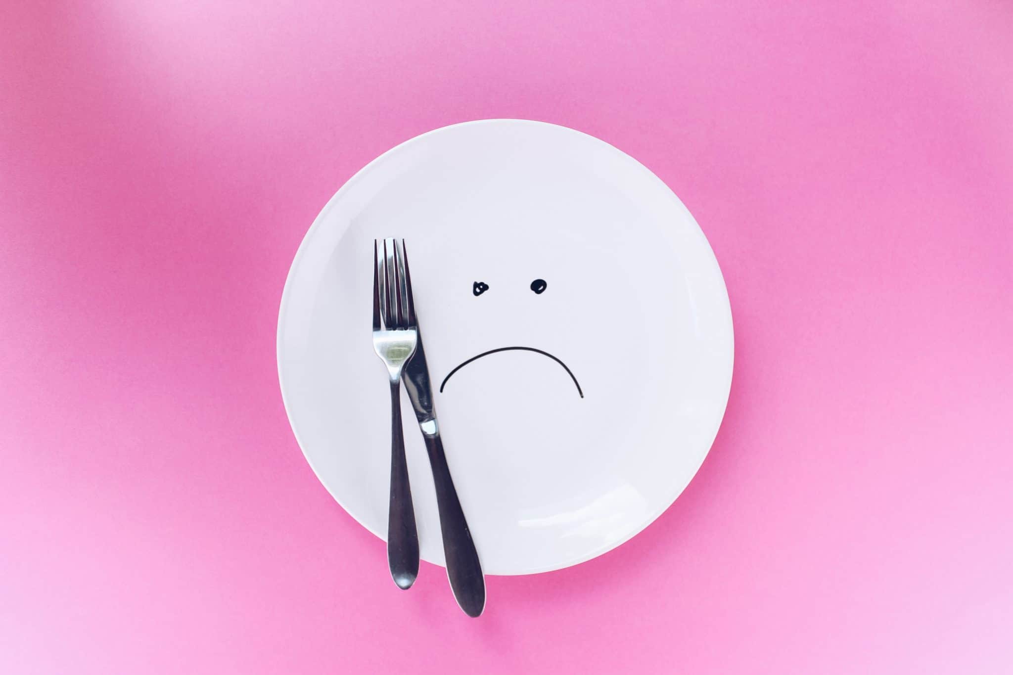 birds eye view of a white plate with a sad face on it with silver cutlery on a pink background