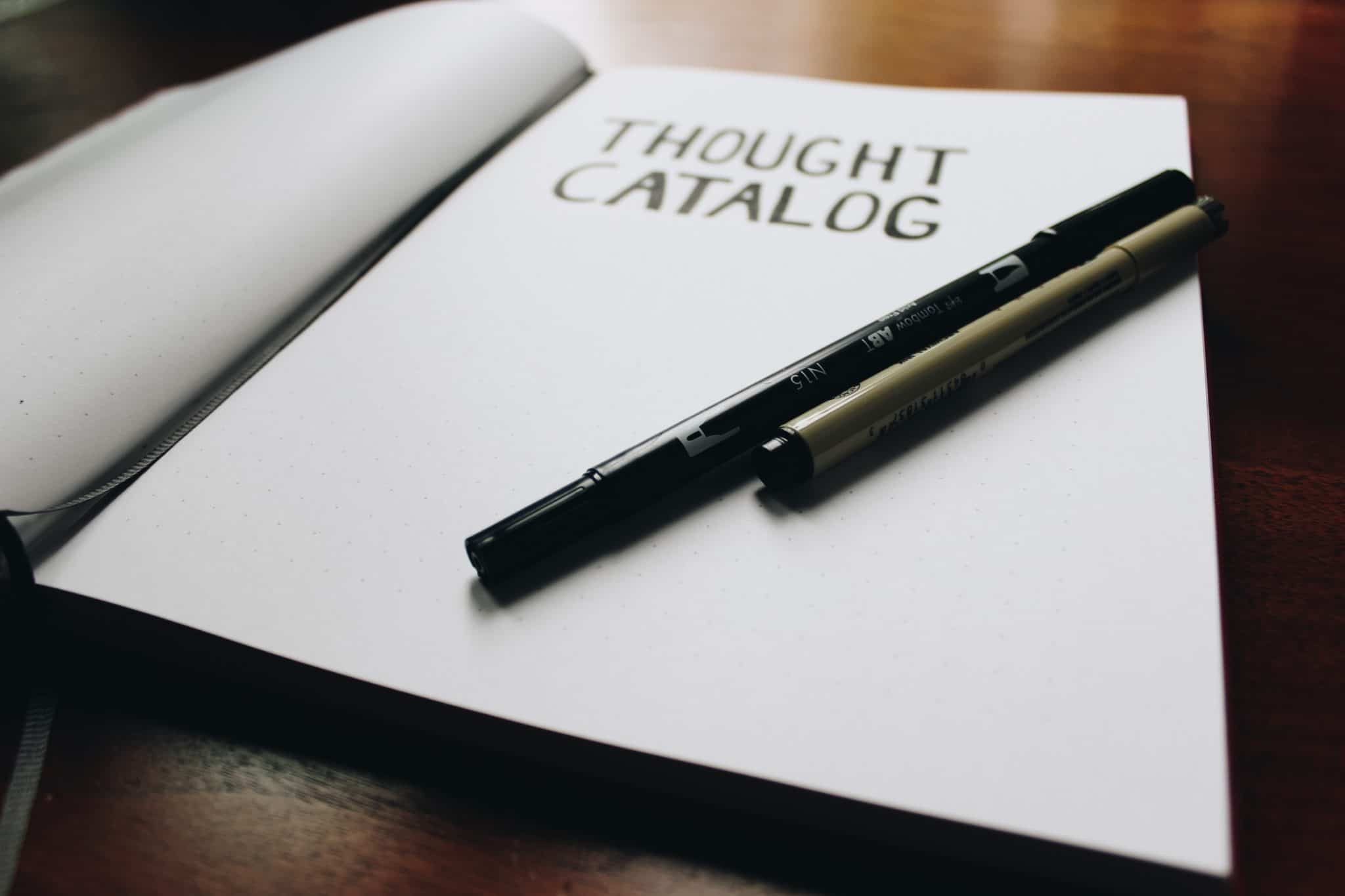 notebook open on a table that reads "thought catalog" next to a black pen