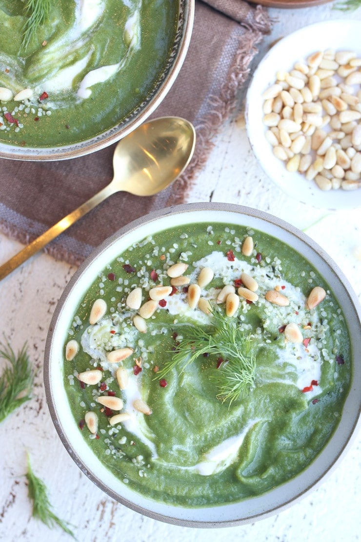Vegan broccoli soup in a bowl with pine nuts, herbs and golden spoon in the background.