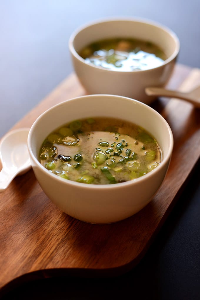 a white bowl of miso soup with green onions and tofu on a brown serving board with a soup spoon beside it.