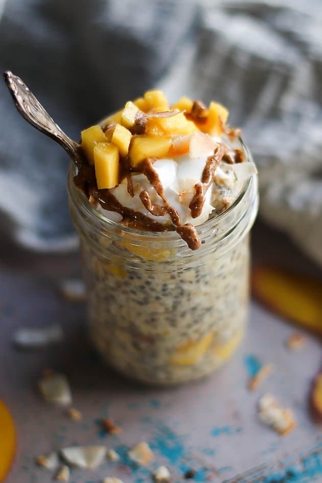 close up of pantry staple mango coconut overnight oats in a glass jar topped with coconut flakes, almond butter, and fresh mango with a silver spoon