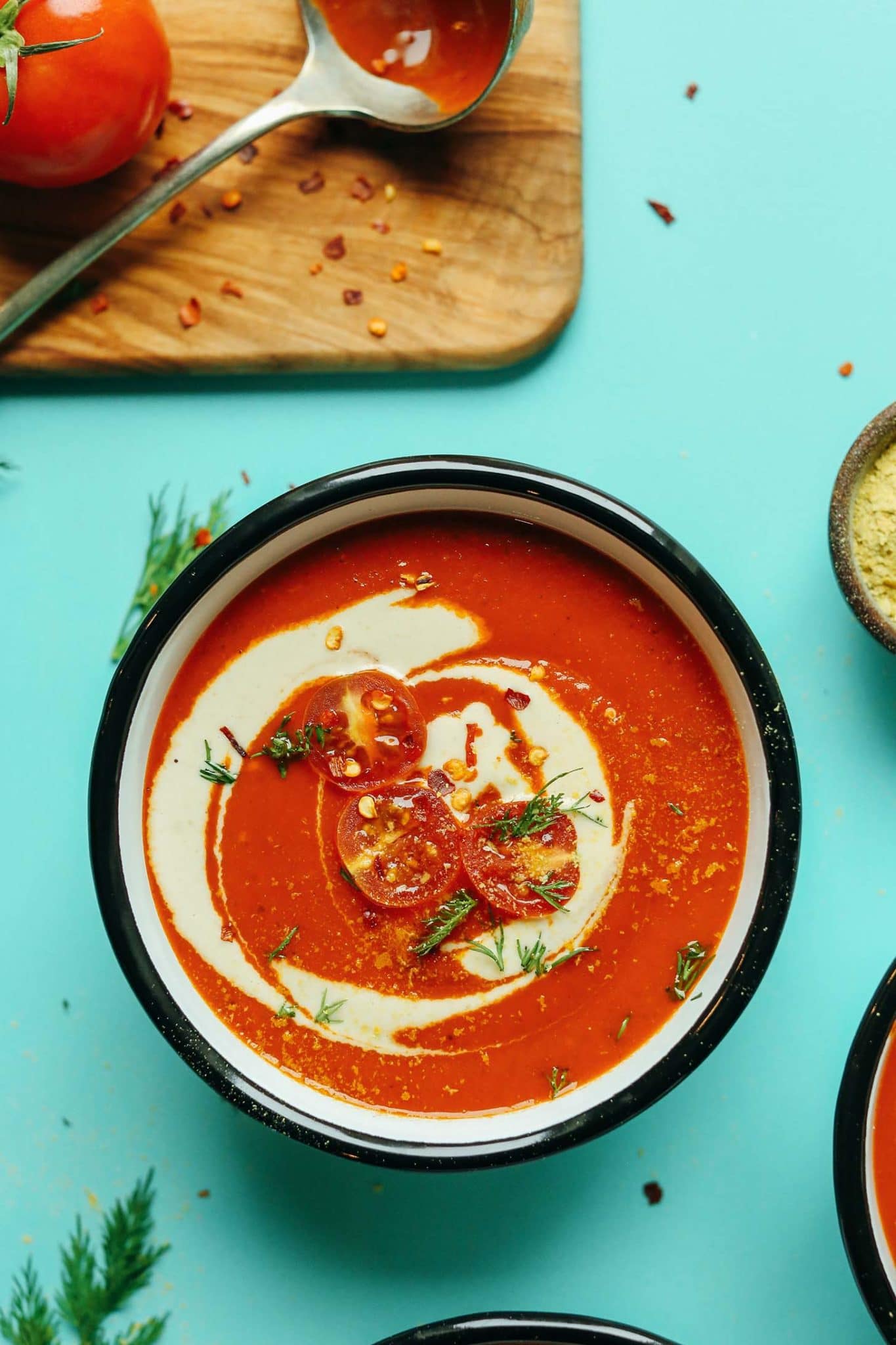 a bowl of creamy roasted red pepper tomato soup topped with tomatoes and greens on a teal background.