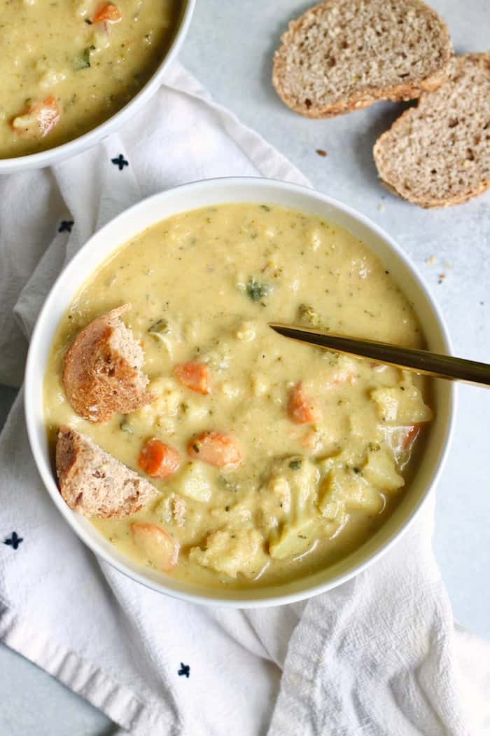 An overhead photo of a white bowl sitting on a white linen with vegan broccoli cauliflower soup inside it topped with some crusty bread.