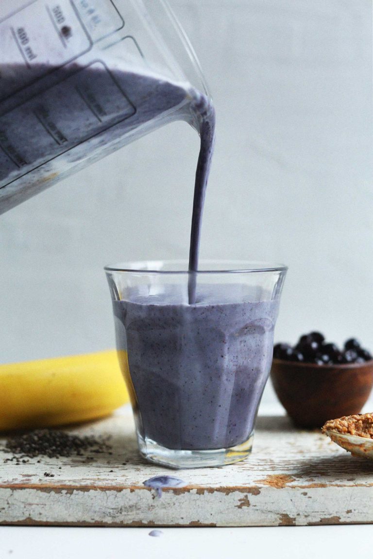 close up of blueberry almond butter smoothie being poured from a blender into a clear glass with a fresh banana and blueberries in the background