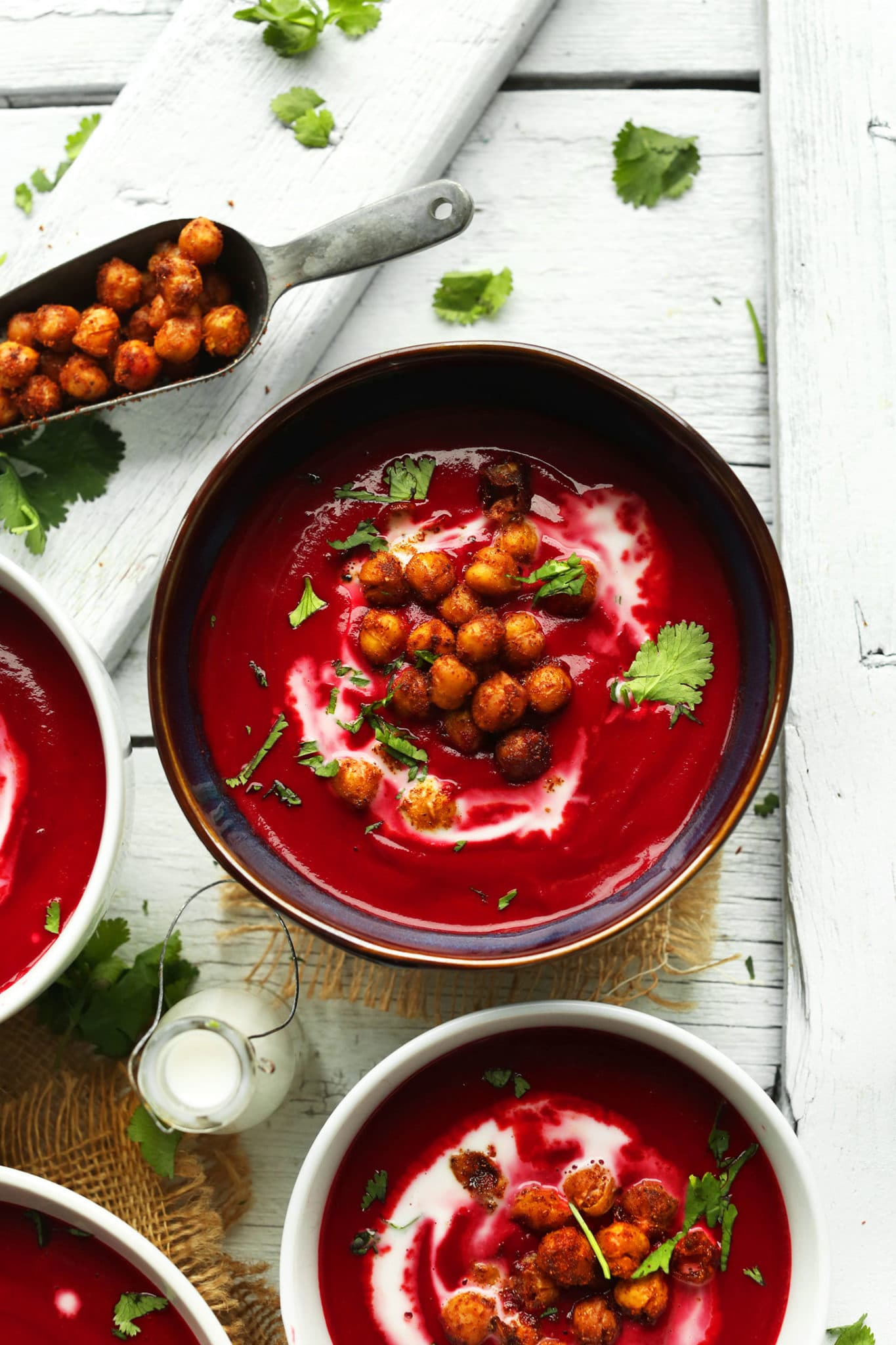 A bowl of curried beet soup topped with tandoori chickpeas and some greens in a dark maroon bowl on a light background with a scoop of chickpeas beside it.