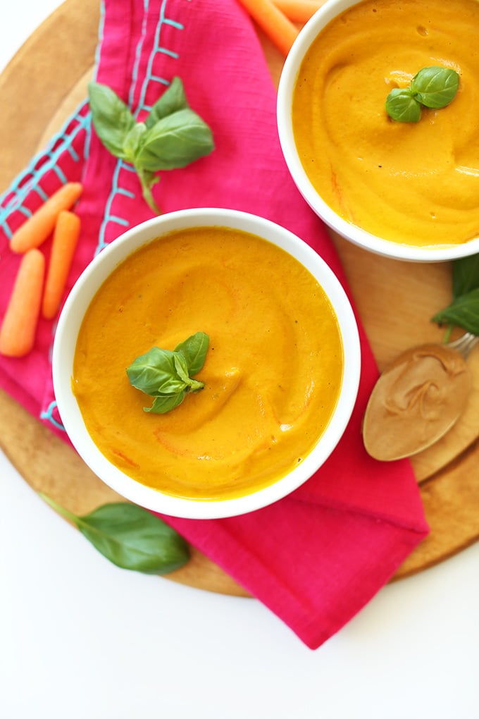 birds eye view of creamy pantry staple thai carrot soup garnished with fresh herbs served on a wooden surface next to fresh carrots and nut butter