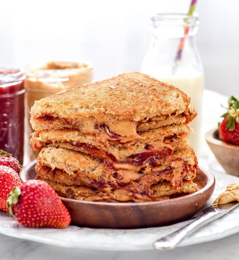 close up of grilled peanut butter and jelly sandwich halves stacked on a plate next to jars of peanut butter and jelly with a glass of milk and fresh strawberries in the background