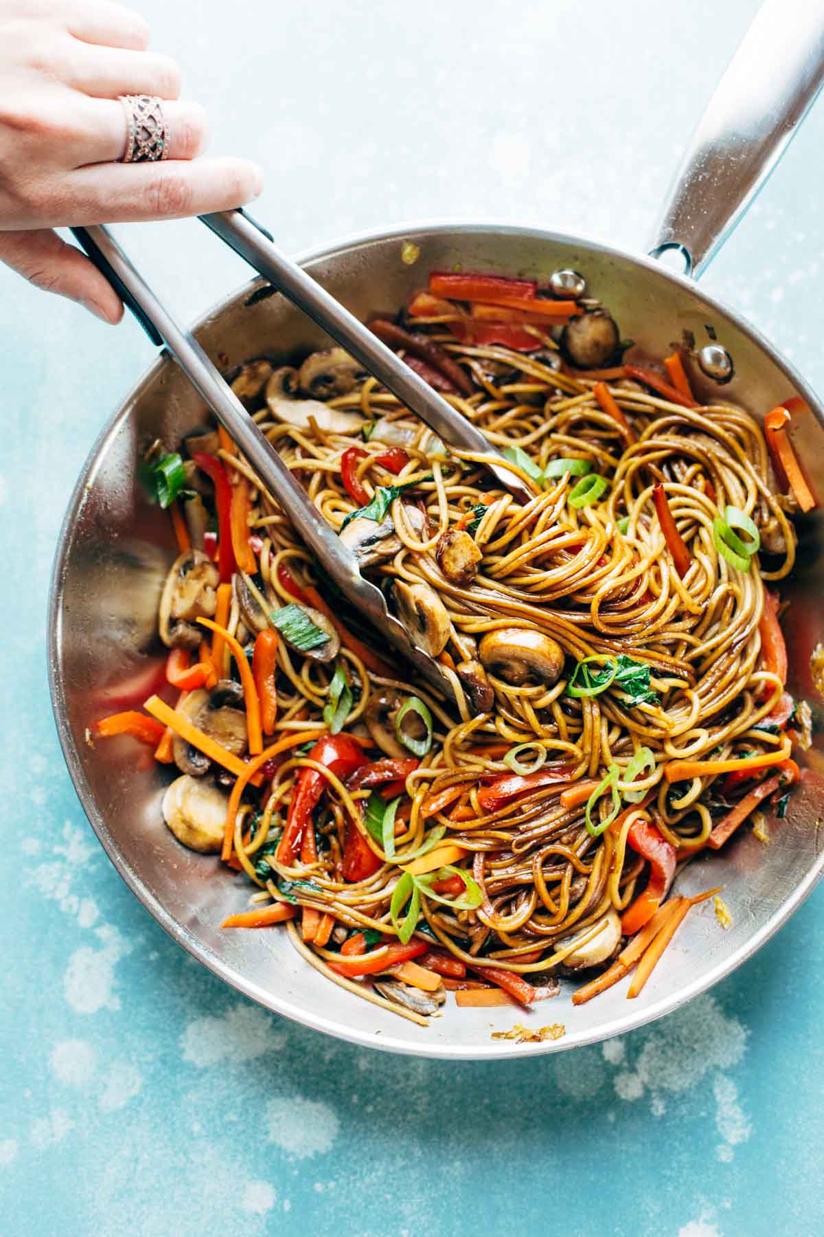 birds eye view of a large silver cooking pan containing fifteen minute lo mein using beans and pulses with tongs inside stirring the noodles