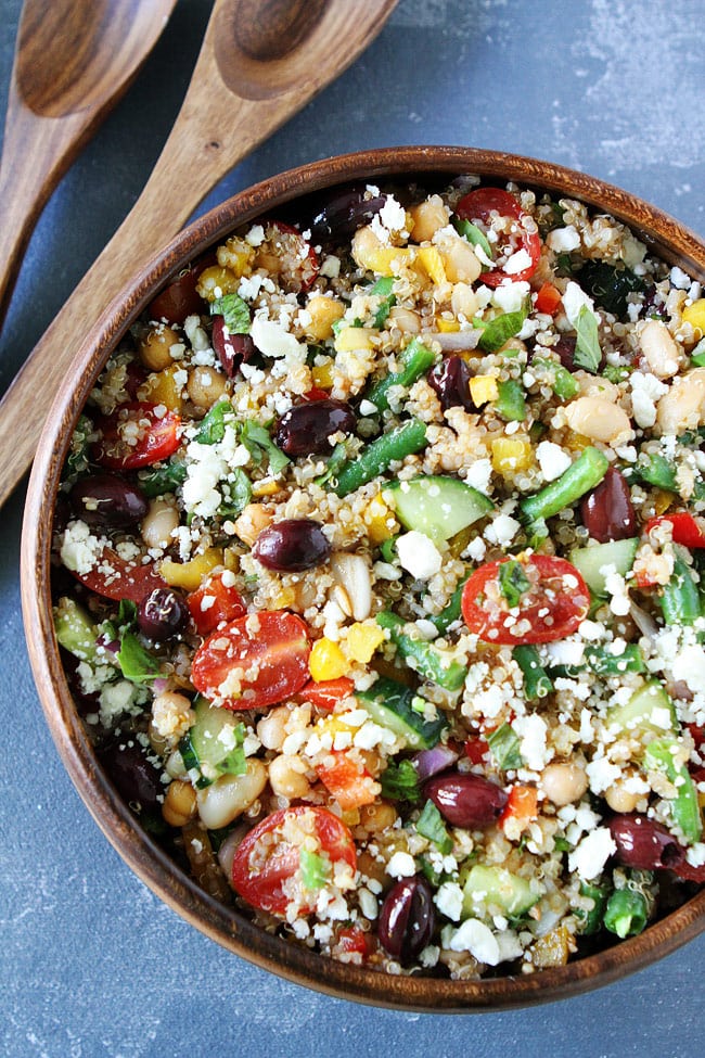 close up of a wooden bowl containing three bean mediterranean quinoa salad garnished with crumbled cheese next to two wooden serving utensils