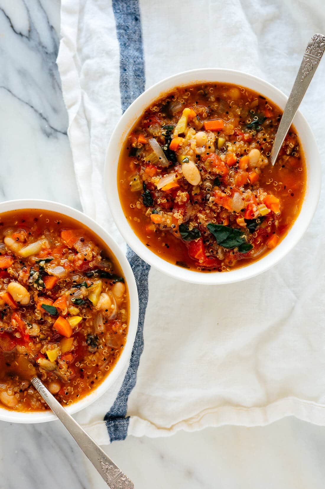 Two bowls of quinoa vegetable soup with kale with a silver spoon inside it on top of a white linen napkin.