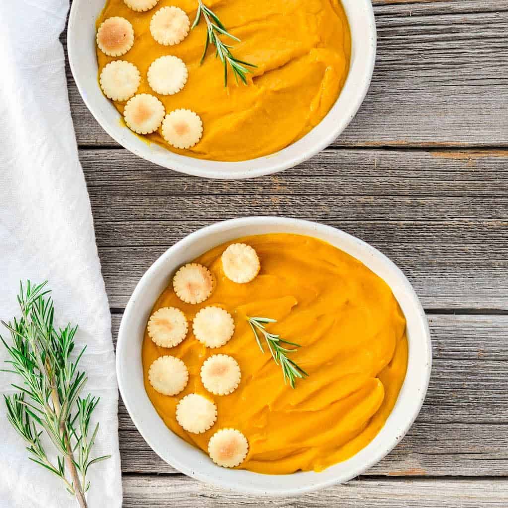 Two bowls of roasted root vegetable soup on a wooden backdrop with rosemary on the side and as a garnish