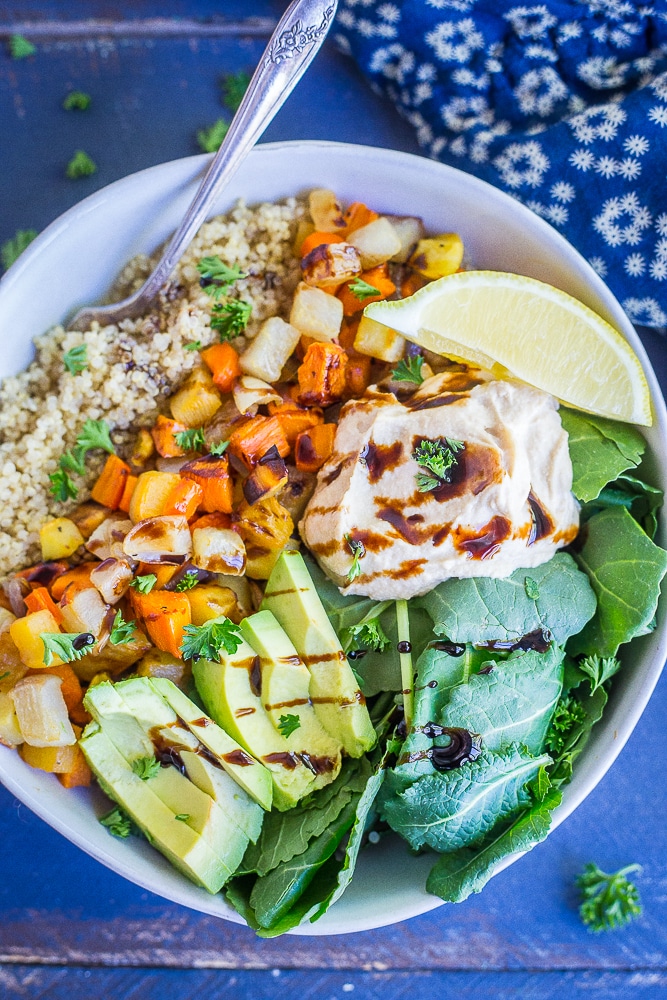 birds eye view of a roasted root vegetable buddha bowl garnished with balsamic, herbs, fresh herbs, and avocado in a white bowl against a blue background