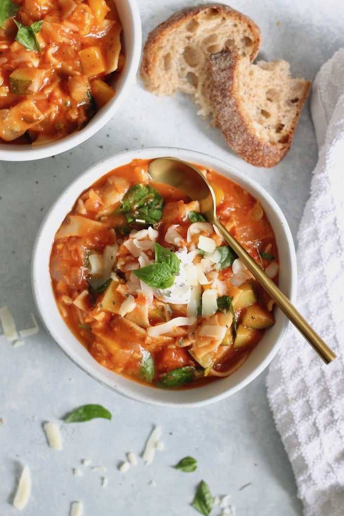 A white bowl on a light background with vegan lasagna soup inside of it with a golden spoon on top.