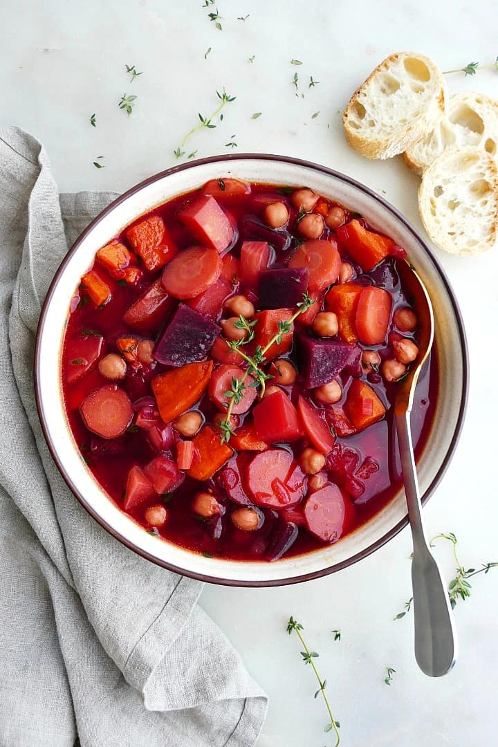 A bowl of vegan stew with winter vegetables in a white bowl with a silver spoon inside of it on a white background with a grey linen napkin beside it.