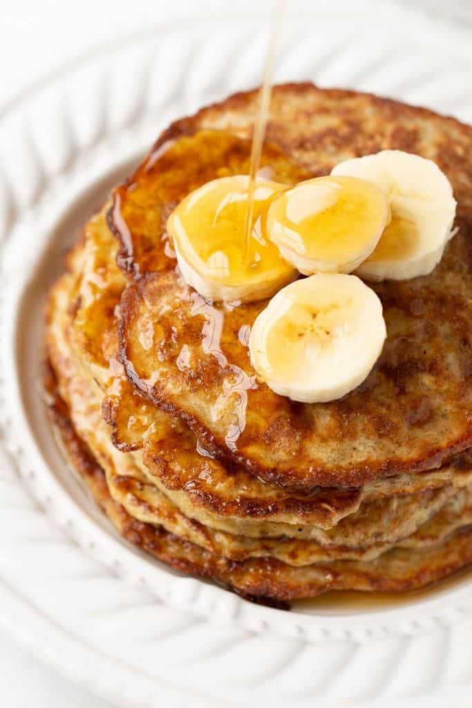 close up of a stack of pantry oatmeal pancakes garnished with banana slices drizzled with maple syrup