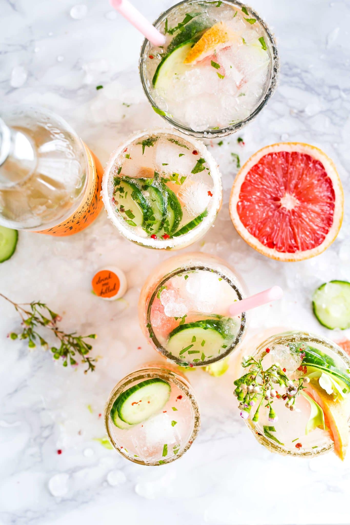 birds eye image of multiple cocktails garnished with fresh herbs, cucumbers, and grapefruit against a white marble background