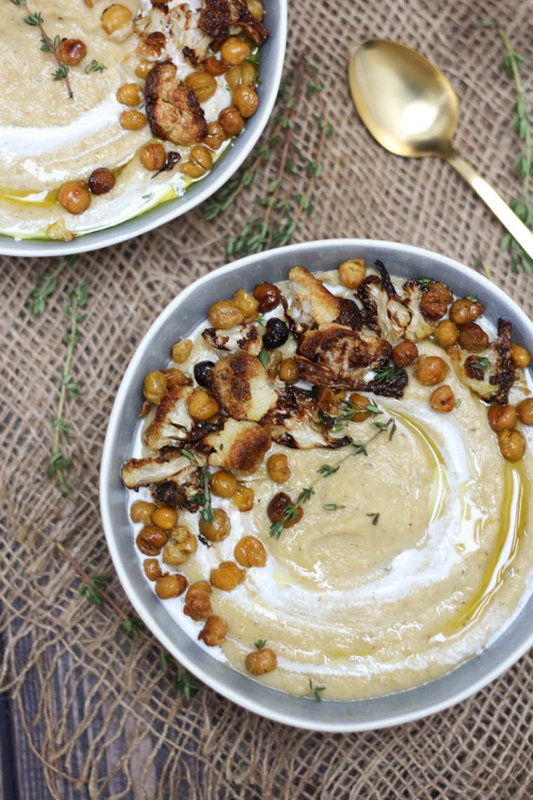 birds eye view of roasted cauliflower soup garnished with chickpeas and herbs in a blue bowl next to a gold spoon and another bowl of soup