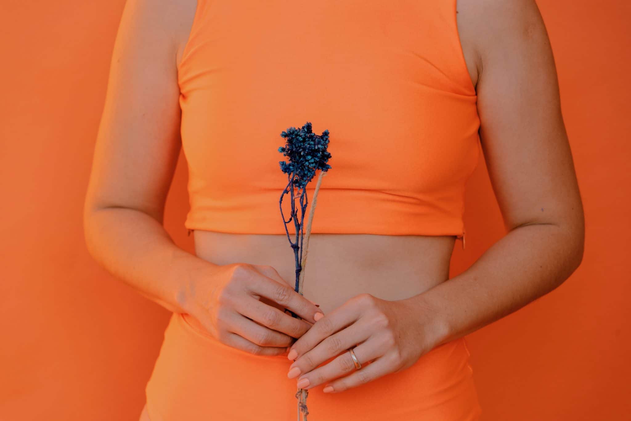 woman standing in orange exercise apparel holding a blue flower against an orange background 