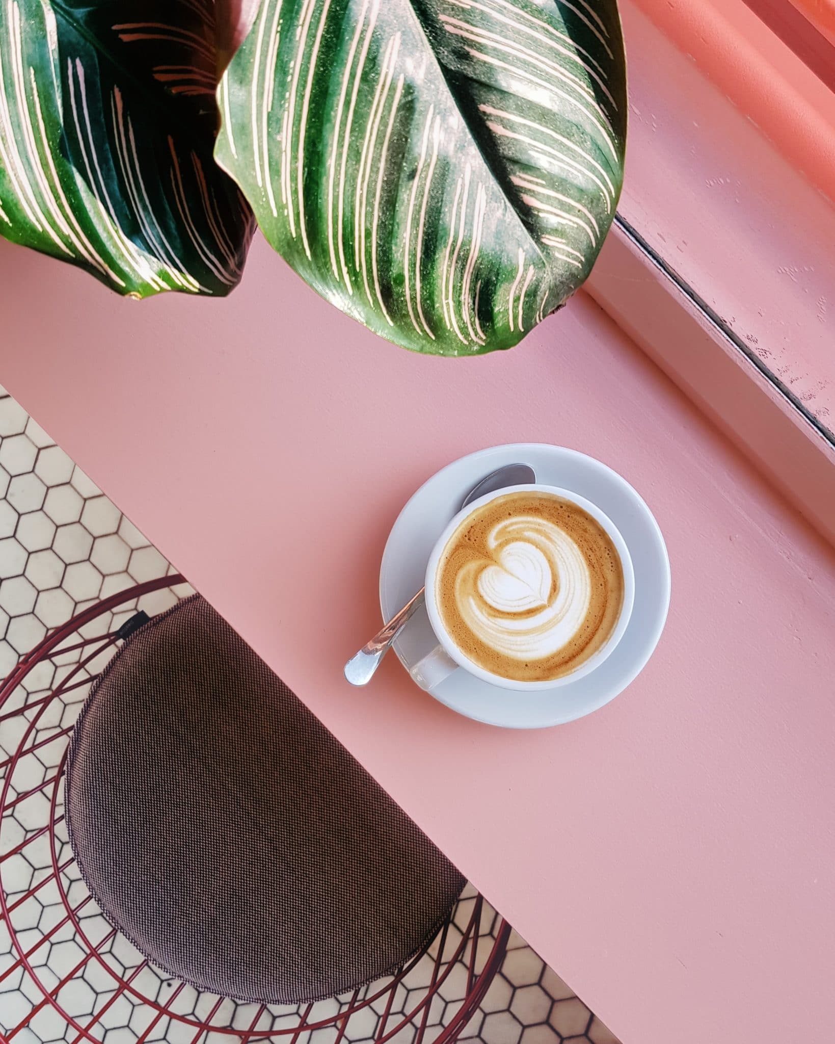 birds eye image of a small mug of coffee on a light pink table next to a grey chair with a green plant in the top left corner