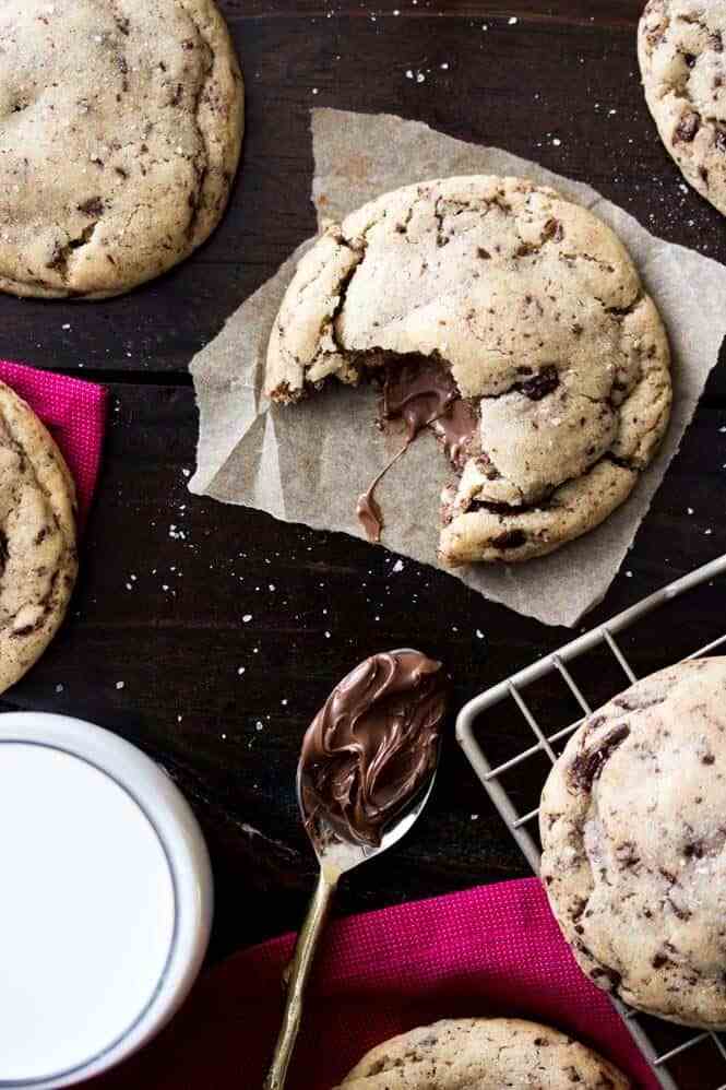 birds eye view image of a bitten into nutella stuffed cookie on a piece of parchment paper next to a spoon of nutella and additional cookies surrounding the parchment paper