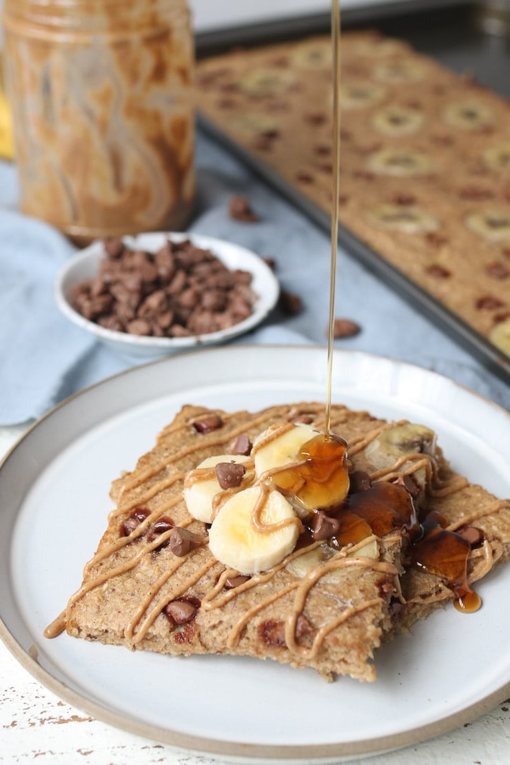 Pancake squares on a white plate topped with bananas and drizzled with maple syrup with the sheet pan in the background. 