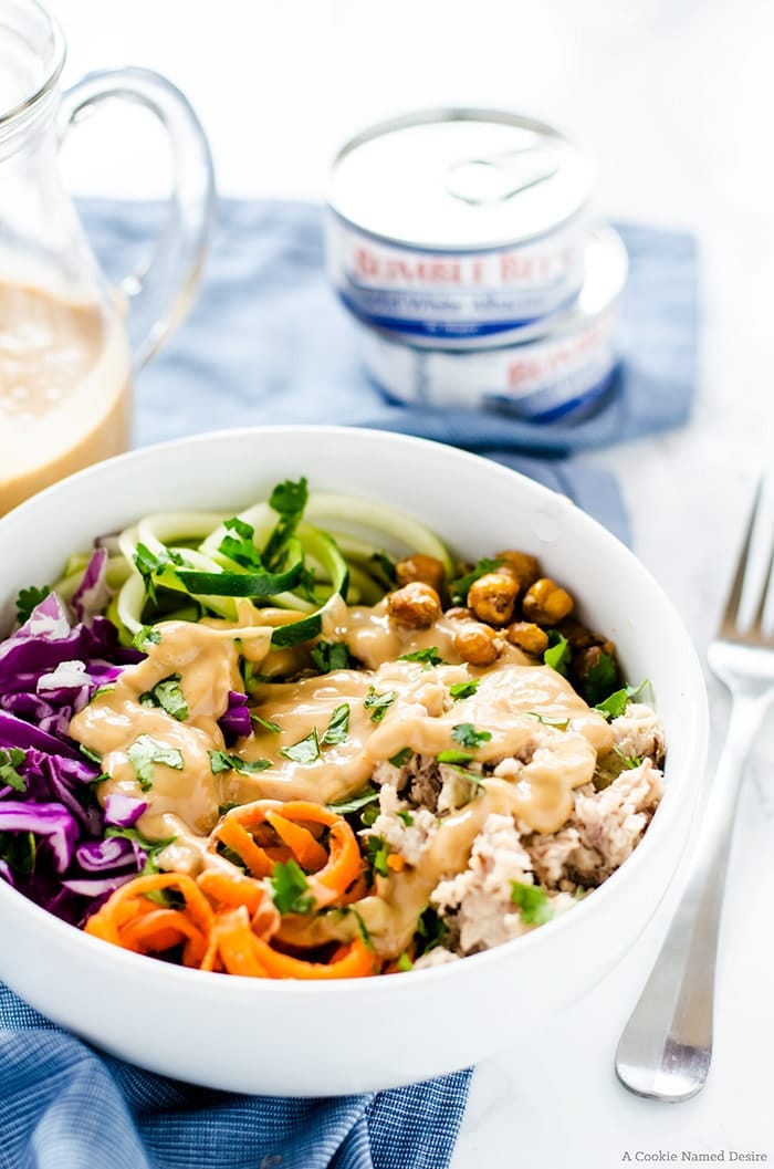 close up of thai tuna power bowl garnished with fresh herbs served in a white bowl with cans of tuna in the background