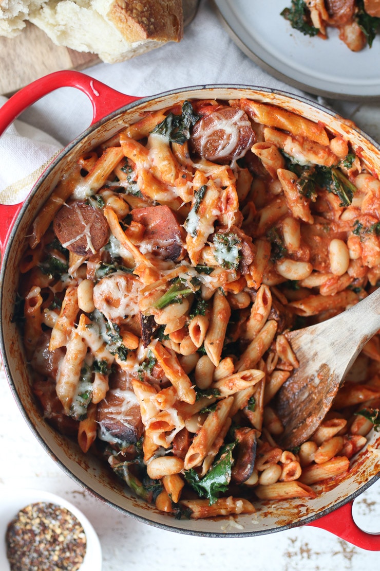 birds eye view of vegan pasta bake with beans and kale in a large red pot with a wooden stirring spoon inside