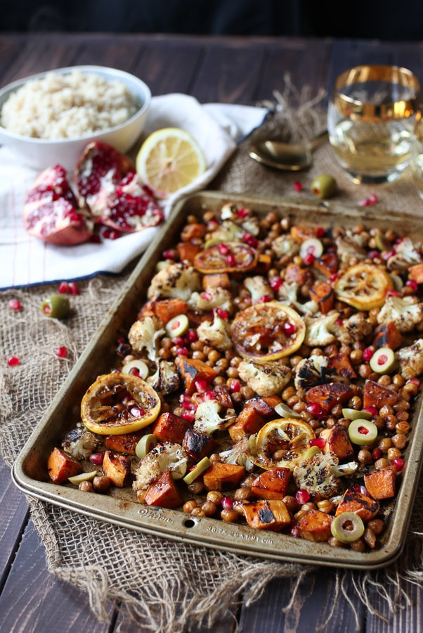 full shot image of chickpea and cauliflower dinner served on a sheet pan with pomegranate seeds, lemon, and cauliflower in the background 