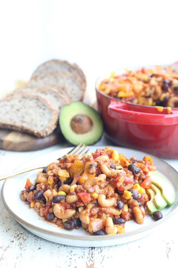 Taco casserole on a white plate with red pot, avocado, and bread in the background