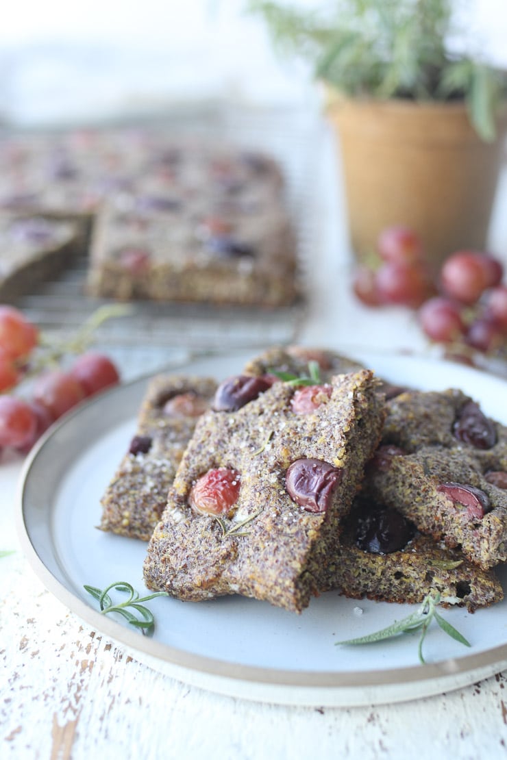 Full shot of gluten free focaccia bread garnished with olives and rosemary on a white dish with additional focaccia and red grapes in the background.