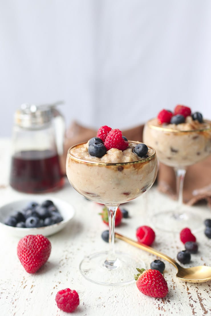 two dessert cups with rice pudding made with oatmeal topped with berries with syrup and blueberries in the background 