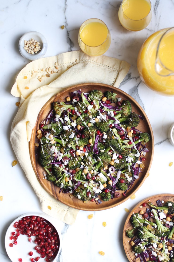 birds eye view of grilled broccoli salad on a brown plate topped with tahini dressing with pomegranate arils, and orange juice pictured beside