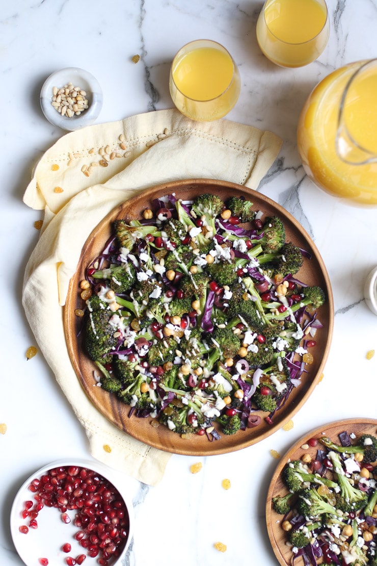 birds eye view of grilled broccoli salad on a brown plate topped with tahini dressing with pomegranate arils, and orange juice pictured beside 