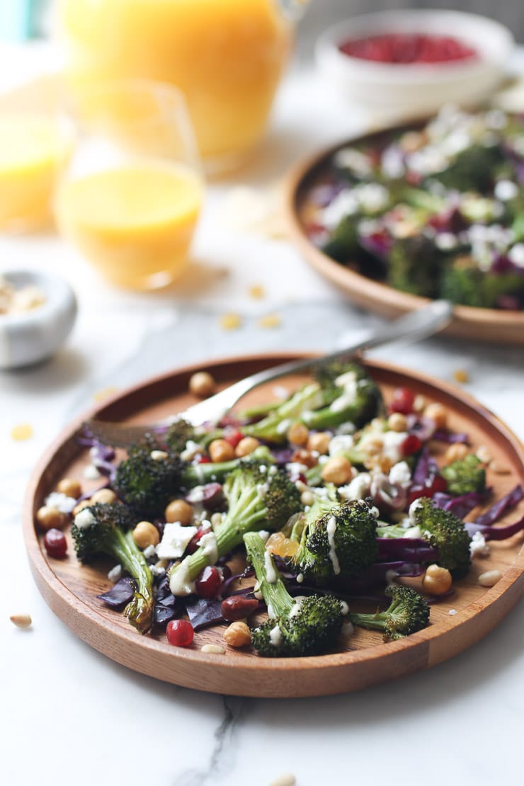 full shot image of vegan roasted broccoli, orange juice, and tahini salad garnished with chickpeas, raisins, pomegranate seeds, pine nuts, and olives on a wooden plate with an additional wooden plate of salad in the background and a clear glass of orange juice