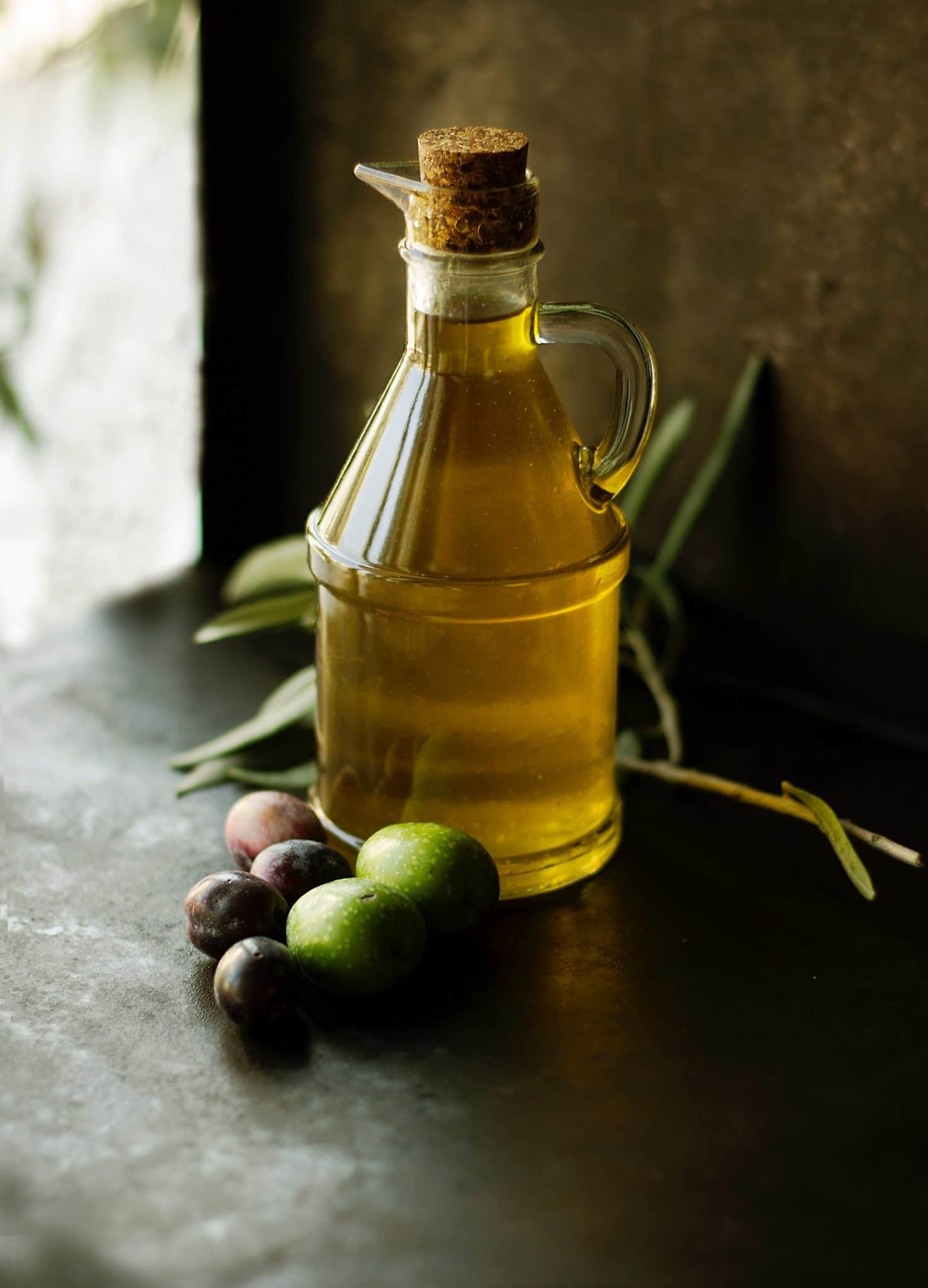 clear jug filled with olive oil on a wooden table next to a small bunch of fresh olives