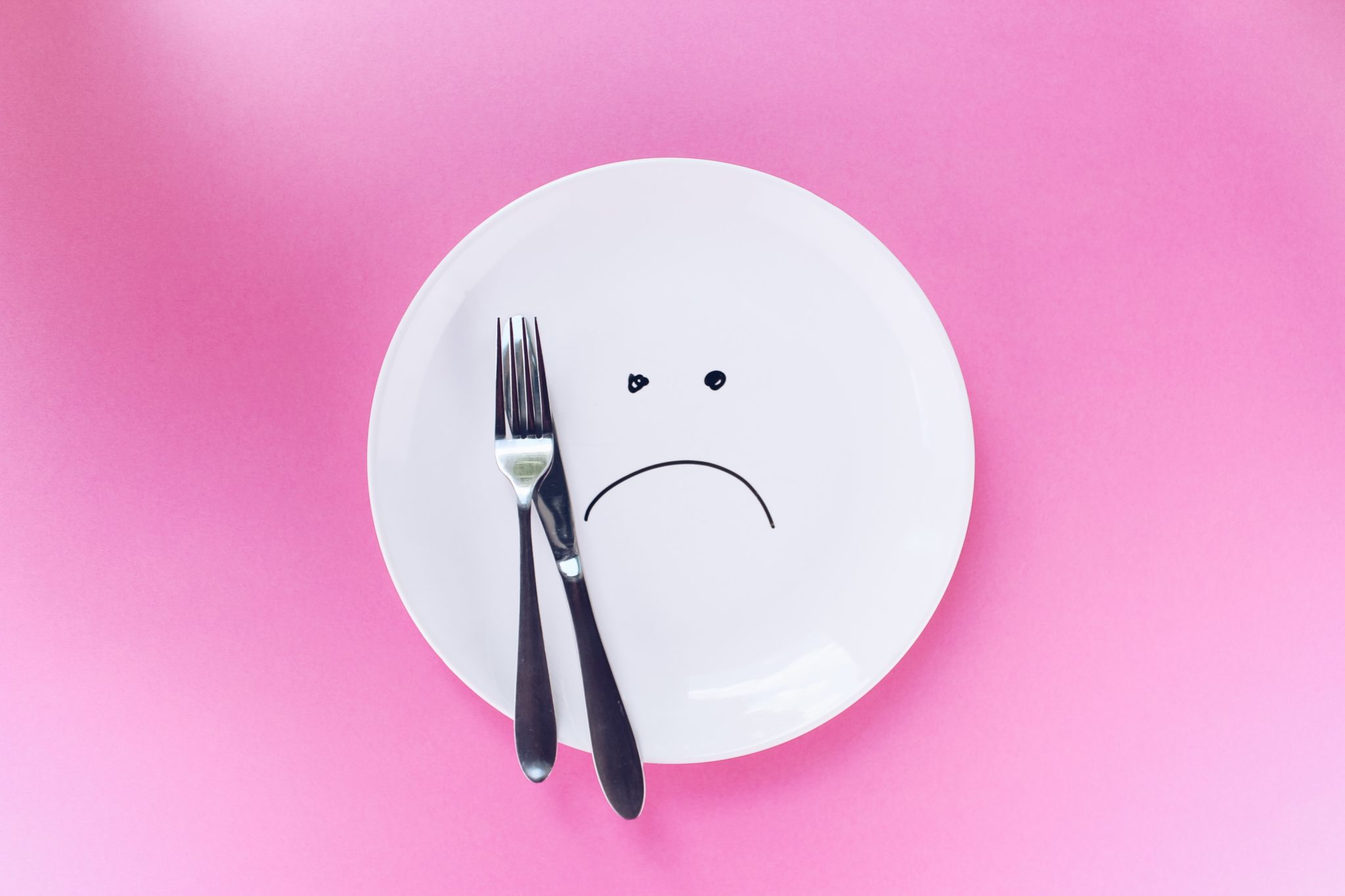 image of a plate with a sad face drawn on top next to a fork and knife against a pink background