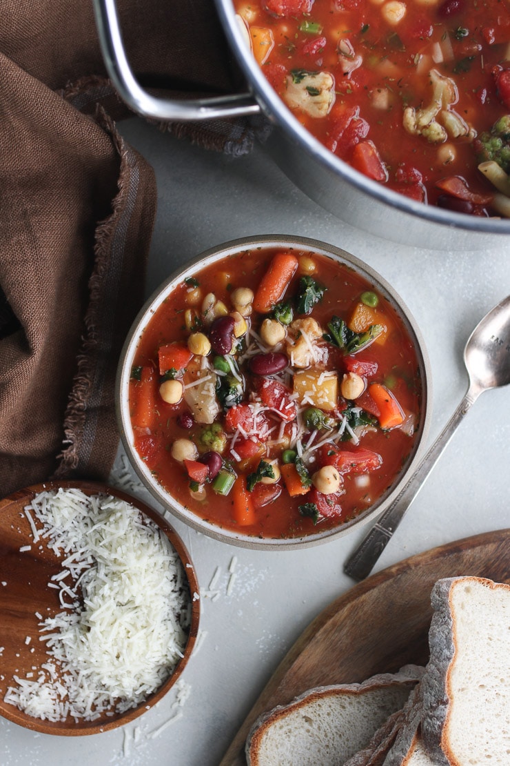 birds eye view of pantry soup recipe with beans & vegetables in a bowl surrounded by vegan cheese, a spoon, bread, and a pot with soup 