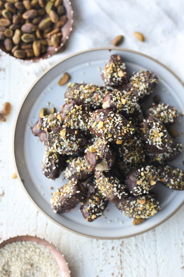 Chocolate covered stuffed dates stacked on a white plate topped with pistachios and sesame seeds.