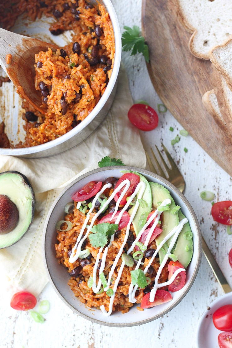 Birds eye view of vegan rice and beans enchilada bowl garnished with cherry tomatoes, fresh herbs, avocado and vegan sour cream against a white background.