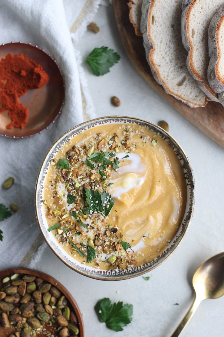 Birds eye view of butternut squash soup with red lentils topped with parsley and nuts with bread, curry paste, and pistachios in the background.